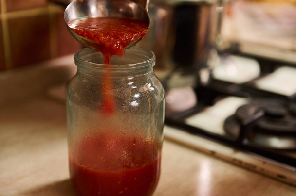 Vista cortada de uma dona de casa em avental de chefs pretos usando uma concha, derrama suco de tomate fervente de panela em jarro esterilizado enquanto cozinha passata caseira na cozinha em casa. Conserva de alimentos - Foto, Imagem