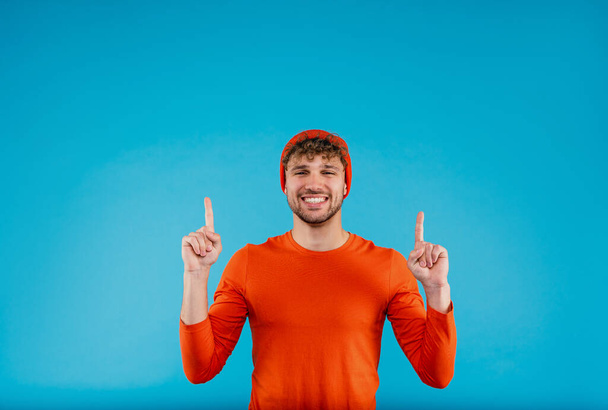 Portrait of young attractive man in orange hat and t-shirt pointing with his fingers up on blue background. High quality photo - Photo, image