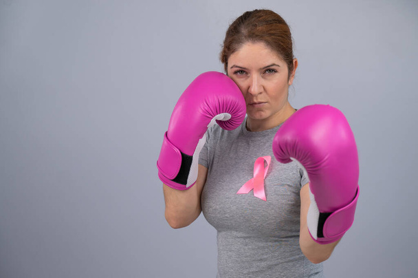 Caucasian woman in pink boxing gloves with a pink ribbon on her chest on a gray background. Fight against breast cancer - Foto, afbeelding