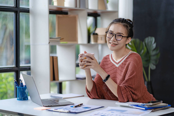 Hymyilevä menestynyt aasialainen nainen työskentelee hänen työpöytänsä ääressä käyttäen tietokoneita ja freelancerien taloudellisia kaavioita. Ecommerce Online Marketing Ideat - Valokuva, kuva