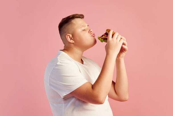 Half-length portrait of cute overweight boy in white t-shirt tasting delicious burger isolated on pink background. Fast food, taste, body positive, emotions and facial expressions. Copy space for ad - Фото, зображення