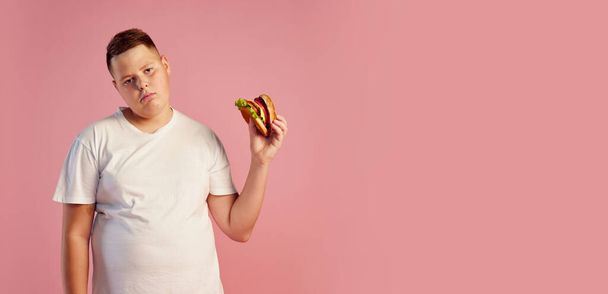 Tired annoyed overweight teen boy in white t-shirt refuses to eat big burger isolated on pink background. Fast food, taste, body positive, dieting and facial expressions. Copy space for ad - Photo, image