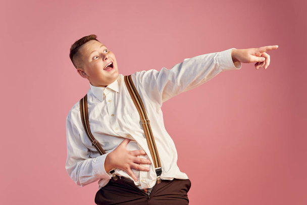 Good sense of humor. Funny teen boy in white shirt and shorts with suspenders laughing isolated over pink background. Concept of health, youth, emotions, retro vintage 70s, 80s american style - Fotó, kép