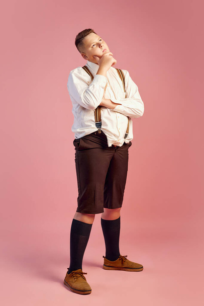 Thoughts, doubts. Cute big obese teen boy in white shirt and shorts with suspenders thinking about life isolated over pink background. Concept of health, youth, emotions, retro vintage 70s, 80s - Zdjęcie, obraz