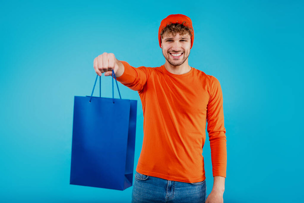 Junger attraktiver Mann hält Einkaufstasche und sieht glücklich und zufrieden aus auf isoliertem blauem Hintergrund. Hochwertiges Foto - Foto, Bild