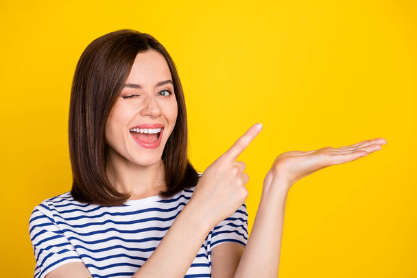 Photo of young attractive smiling cute woman blinking finger directing holding palm empty space advert isolated on yellow color background. - Photo, image