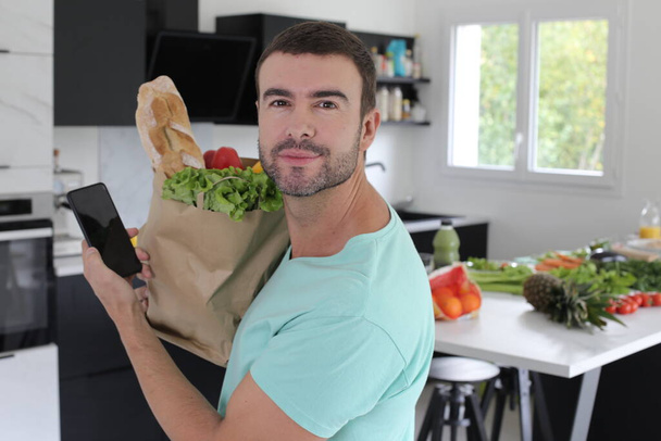 portrait of handsome young man holding paper bag of groceries at kitchen - Photo, Image
