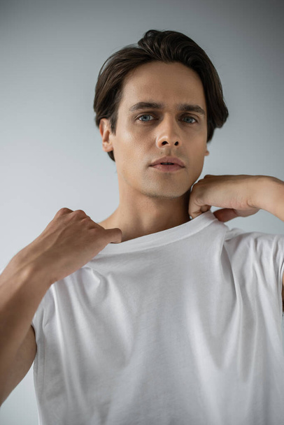 homme élégant regardant la caméra et ajustant t-shirt blanc sur gris - Photo, image
