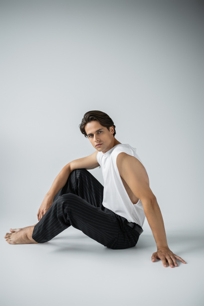 barefoot man in trousers and white t-shirt looking at camera while sitting on grey  - Foto, Imagem