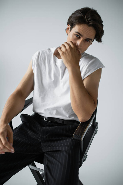 brunette man in striped trousers and white t-shirt sitting on camp chair on grey  - Foto, immagini