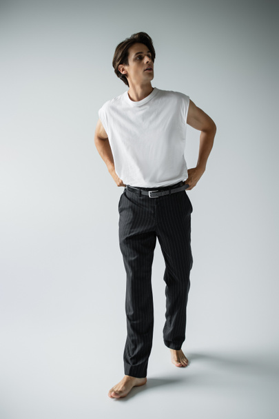 full length of barefoot man in white t-shirt and black pants posing while standing on grey - Fotografie, Obrázek