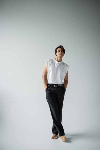 full length of stylish man in white t-shirt and black pants posing while standing with crossed legs on grey - Fotoğraf, Görsel
