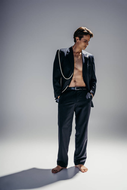 full length of brunette man with hairy chest and stylish suit with pearl chain posing with hands in pockets on dark grey - Foto, Imagem