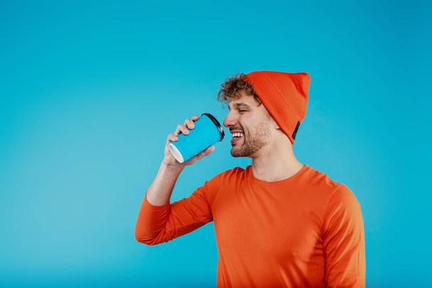 Yound attractive happy man in orange hat and t-shirt holds paper cup with coffee tea on blue background. High quality photo - Valokuva, kuva