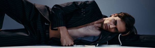 stylový a brunetka muž v obleku s perlami ležící při pózování na tmavě šedé, prapor - Fotografie, Obrázek
