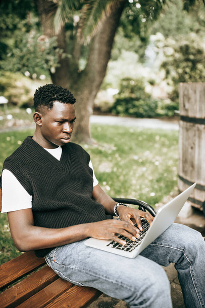 Jonge zwarte man in vest werkt buiten op laptop computer, serieus of bezorgd kijkend naar het scherm. Studenten zitten op een bank op de universiteitscampus, doen freelance afstandsonderwijs - Foto, afbeelding
