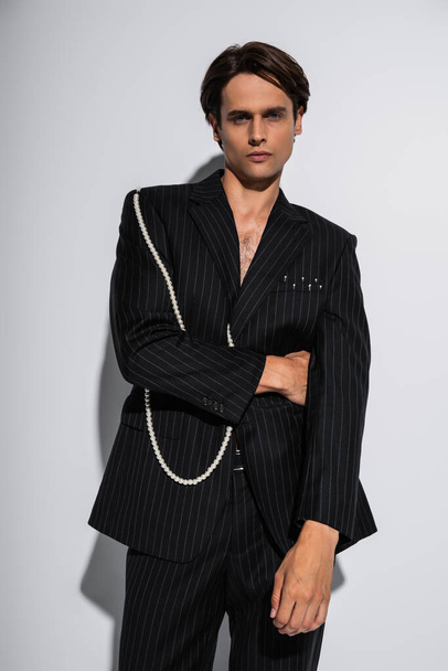 brunette man in trendy suit with pearls posing while looking at camera on grey - Foto, Imagem