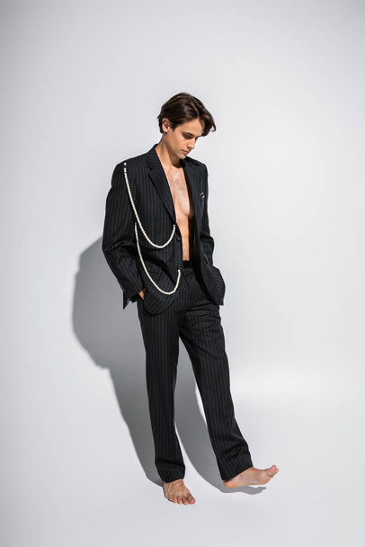 full length of barefoot and brunette man in trendy suit with pearls standing with hands in pockets on grey  - Fotografie, Obrázek