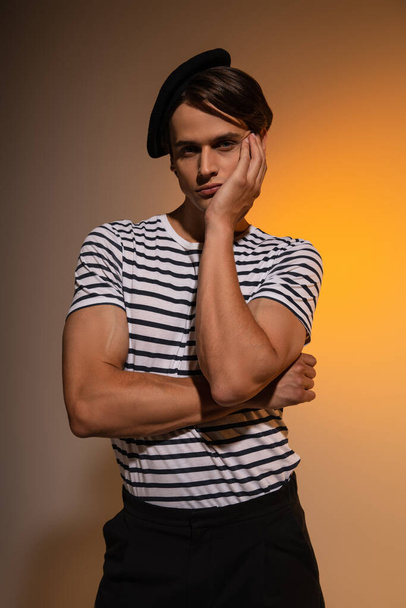 stylish man in beret and striped t-shirt looking at camera while posing on orange  - Fotografie, Obrázek