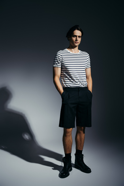 Full length of trendy man in striped t-shirt and shorts holding hands in pockets on grey background with lighting  - Фото, зображення
