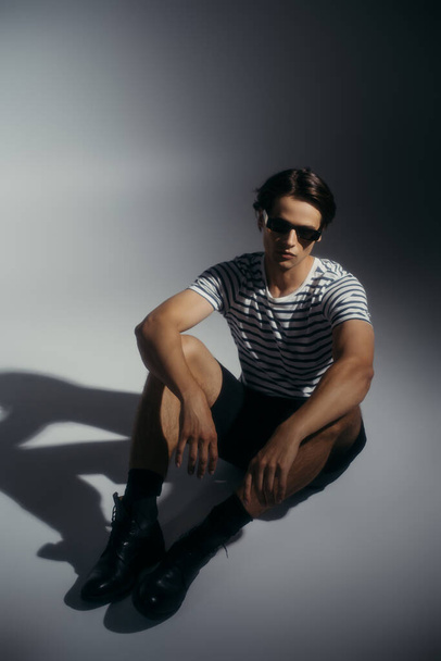 Man in sunglasses and striped t-shirt sitting on grey background with shadow  - Foto, immagini