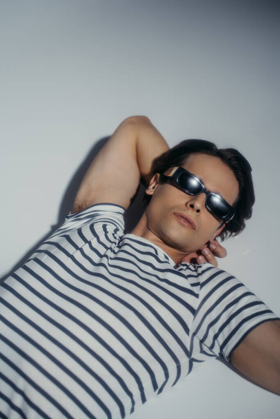 High angle view of stylish man in sunglasses and striped t-shirt lying on grey background - Foto, Imagem