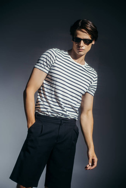Modelo en gafas de sol y pantalones cortos posando sobre fondo gris con iluminación  - Foto, imagen