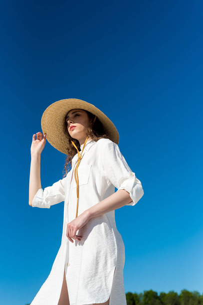 młoda piękna kobieta w białej sukience i kapeluszu pozowanie na tle niebieskiego nieba - Zdjęcie, obraz