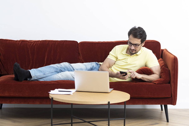 Beau homme allongé sur le canapé et travaillant sur un ordinateur portable à la maison. Éducation en ligne et concept de travail à distance - Photo, image