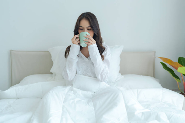 Όμορφη κοπέλα που κάθεται και πίνει ζεστό τσάι το πρωί σε ένα μαλακό λευκό κρεβάτι Νιώστε χαλαροί κάτω από τις ζεστές κουβέρτες. - Φωτογραφία, εικόνα