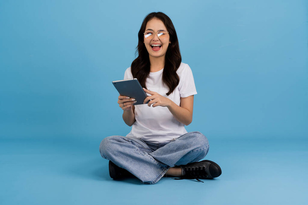 Bella ragazza felice tenendo tablet computer seduto su sfondo blu Siediti e sorridi con una postura rilassata Goditi la navigazione sul web con dispositivi moderni. - Foto, immagini