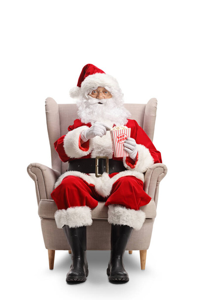 Kerstman zitten in fauteuil en het eten van popcorn geïsoleerd op witte achtergrond - Foto, afbeelding