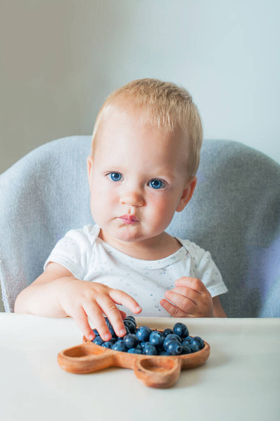 Blondynka maluch chłopiec jedzenie pyszne jagody na wysokim krześle zbliżenie i skopiować przestrzeń. - Zdjęcie, obraz