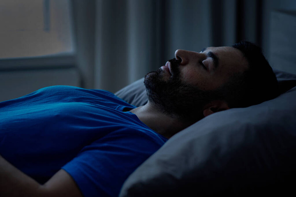 Un hombre milenario relajado de apariencia oriental duerme acostado con los ojos cerrados en el dormitorio de su casa. Concepto de sueño saludable. Vista lateral. Foto de alta calidad - Foto, imagen