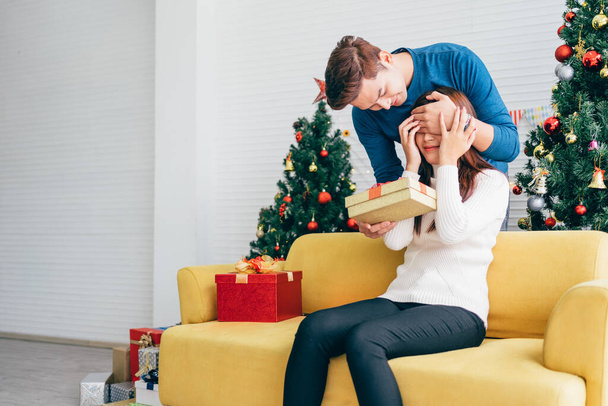 Junger asiatischer Mann überrascht und schließt ihre Augen mit einem Weihnachtsgeschenk zu Hause mit einem Weihnachtsbaum im Hintergrund. Bild mit Kopierraum. - Foto, Bild