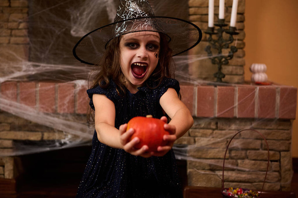 Urocza dziewczynka w czarodziejskiej sukience i czarodziejskim kapeluszu z makijażem artystycznym na twarzy, śmieje się trzymając dynię przed kamerą, siedząc przy kominku pokrytym pajęczynami, w gotyckim wnętrzu domu. Halloween - Zdjęcie, obraz