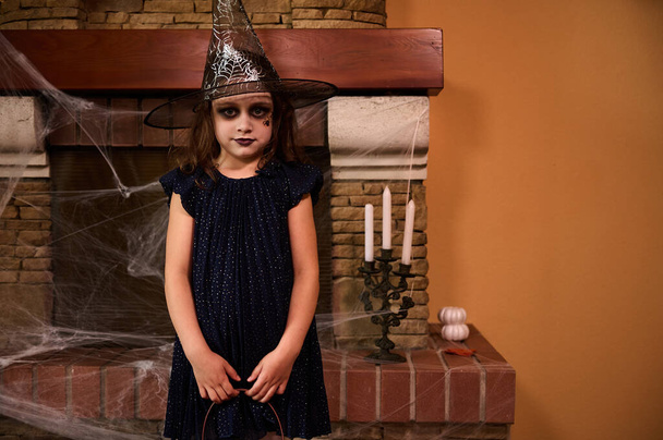 Mischievous mysterious Caucasian little child girl in witch costume and wizard hat, stands against a cobweb covered fireplace and candles, ready for Halloween party. Halloween trick or treat - Photo, Image