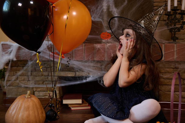 Έκπληκτο λευκό κοριτσάκι, ντυμένο με καπέλο μάγου και μάγου, κοιτάζοντας πορτοκαλί μαύρα μπαλόνια ενώ καθόταν πάνω σε ένα πλακόστρωτο τζάκι. Αποκριάτικο πάρτι. Γοτθική γιορτή. 31 Οκτωβρίου - Φωτογραφία, εικόνα