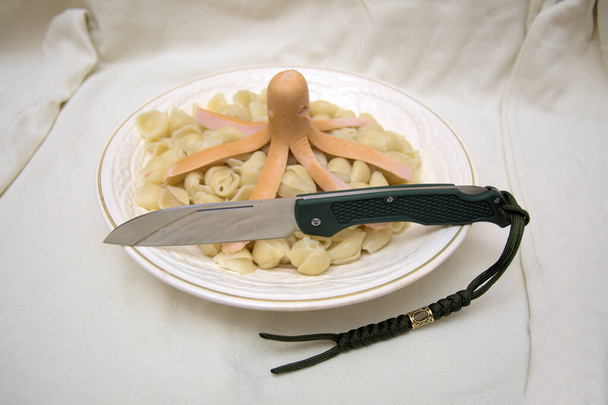 Gourmet dinner octopus sausage with pasta white plate folding knife cutting edge green handle lanyard para cord humor macro background - Photo, Image