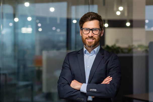 Portrait of successful mature boss, senior businessman in glasses and business suit looking at camera and smiling, man with crossed arms working inside modern office building. - Foto, immagini