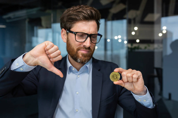 Close-up photo of male businessman holding gold crypto currency bitcoin coin in hands, mature investor disappointed with money rate and investment, boss in business suit showing finger down - Foto, imagen