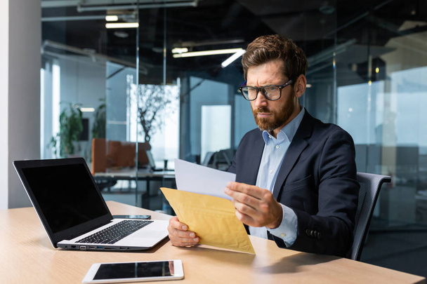 Serious and focused mature boss working inside modern office building with laptop, businessman in business suit carefully reading letter from bank, upset and disappointed man in glasses and beard. - Foto, Imagem