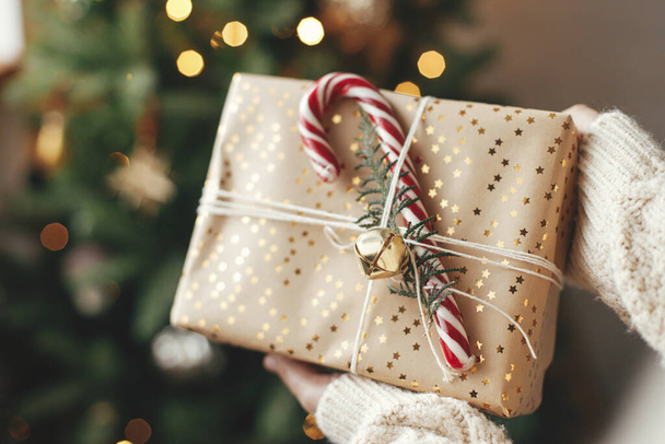 Stilvolles Weihnachtsgeschenk in den Händen gegen Weihnachtsbaum mit Lichtern. Frohe Weihnachten und frohe Feiertage! Frau im kuscheligen Pullover hält mit Zuckerrohr umwickeltes Geschenk in stimmungsvollem Raum - Foto, Bild