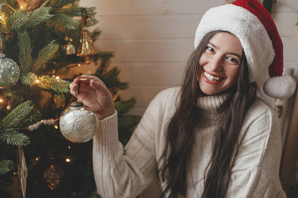 Merry Christmas! Happy woman in cozy sweater and santa hat decorating christmas tree with stylish baubles in atmospheric festive room. Winter holidays preparation - Foto, Imagem