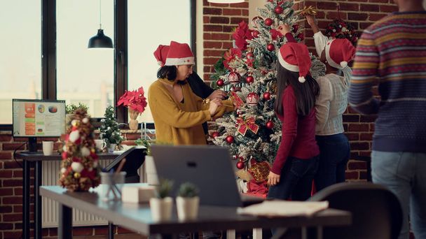 Különböző kollégák díszítéseket tesznek karácsonyfára, megünneplik a téli ünnepeket a munkahelyen. Díszítő iroda karácsonyi fények és ünnepi díszek, érzés boldog szezonális dekoráció. - Fotó, kép
