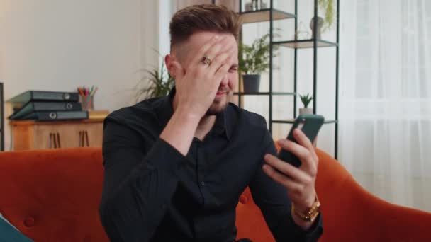 Epäonninen nuori parrakas mies käyttää älypuhelimen kirjoittamista selailu, menettää tulossa yllättynyt äkillinen arpajaiset tuloksia, huonoja uutisia, onni menetys, epäonnistua. Liikemies kaveri kotona olohuoneessa istuu oranssi sohva - Materiaali, video