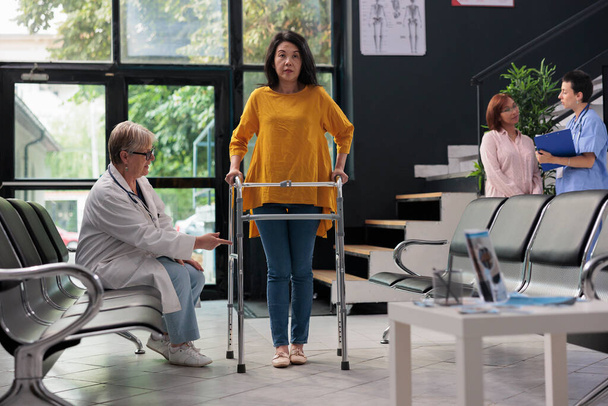 病院のロビーで理学療法で歩行フレームを使用して、事故から回復するために足の骨折を持つアジアの女性を助ける高齢者医療。外傷を負った患者を支援する医師. - 写真・画像