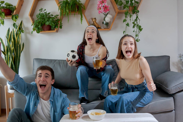 Три молодых друга, две женщины и мужчина, с эйфорией и радостью кричат, смотрят футбольный матч. - Фото, изображение
