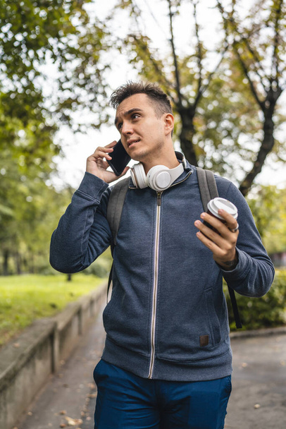 One young adult caucasian man walking in the city town near park using mobile phone smartphone talk with cup of coffee in autumn or spring day happy male tourist standing alone real people copy space - Фото, изображение