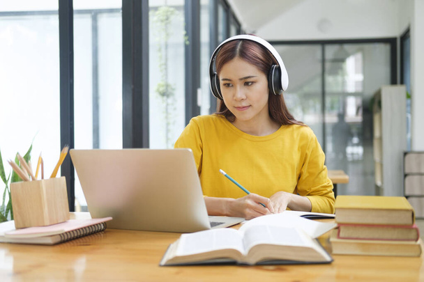 若いアジアの女性学生は、オンラインコースを学び、机に座りながらノートを書き、ヘッドフォンを装着したラップトップや教科書を使用して眼鏡をかけています。オンライン学習と学習の概念. - 写真・画像
